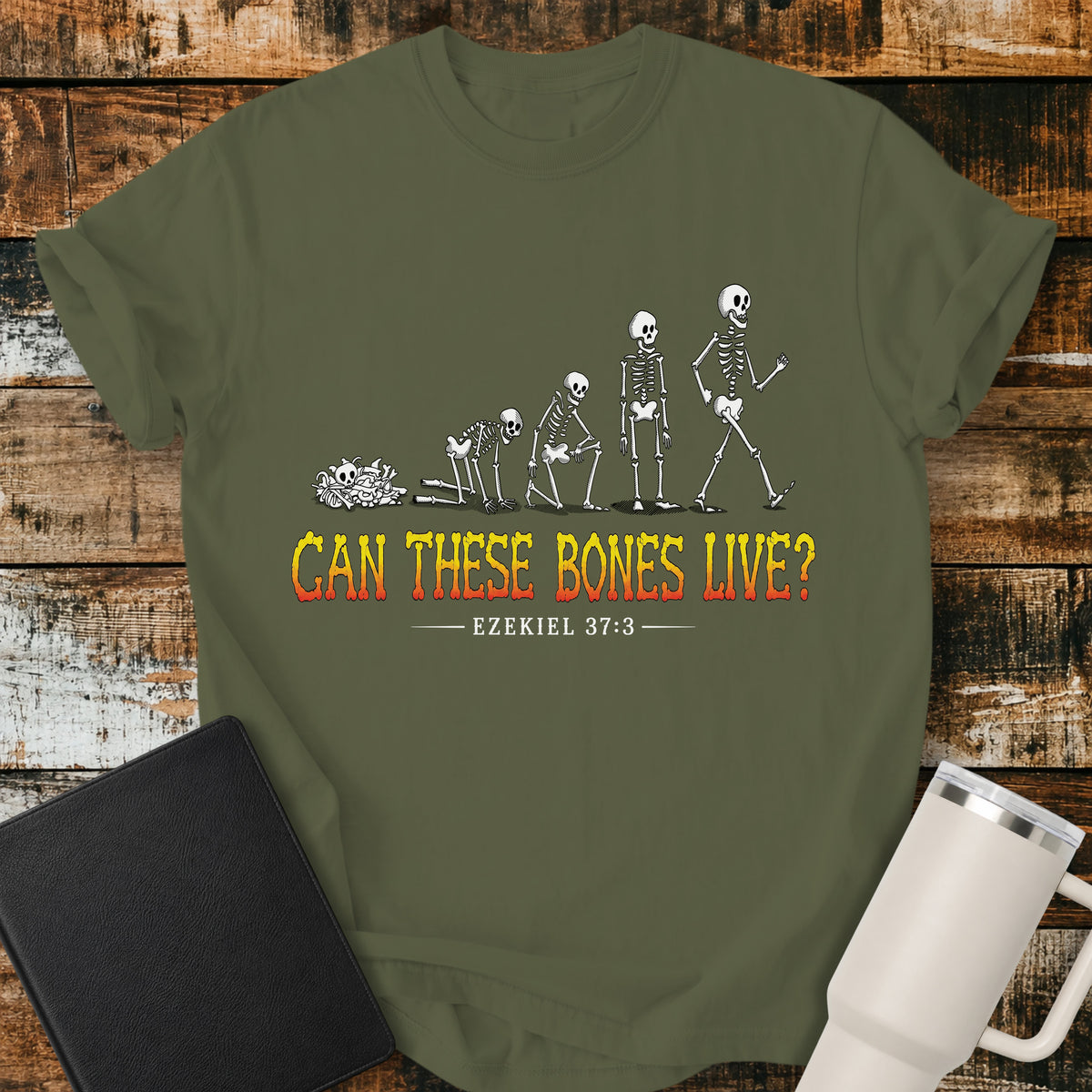 hemp green flat-lay t-shirt mockup featuring a striking illustration of dry bones transforming into a walking skeleton, inspired by Ezekiel's vision of the valley of dry bones. Bold distressed lettering in a yellow-to-red gradient reads, "CAN THESE BONES LIVE?" with the Bible reference "EZEKIEL 37:3" below. The shirt is displayed on a rustic wood plank background alongside a black leather Bible and a white insulated travel mug.
