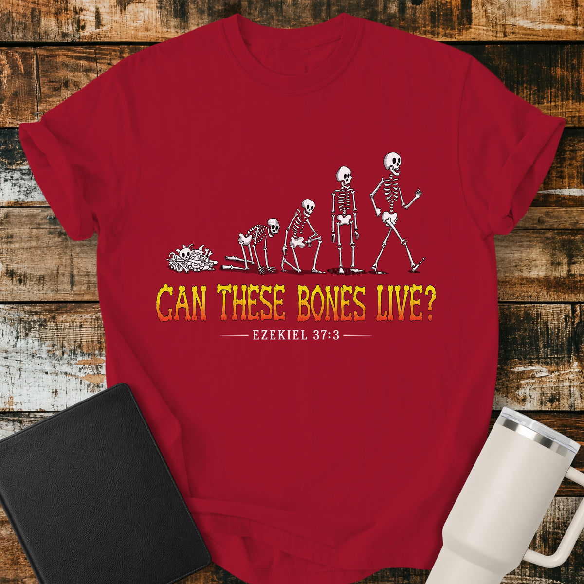 red flat-lay t-shirt mockup featuring a striking illustration of dry bones transforming into a walking skeleton, inspired by Ezekiel's vision of the valley of dry bones. Bold distressed lettering in a yellow-to-red gradient reads, "CAN THESE BONES LIVE?" with the Bible reference "EZEKIEL 37:3" below. The shirt is displayed on a rustic wood plank background alongside a black leather Bible and a white insulated travel mug.