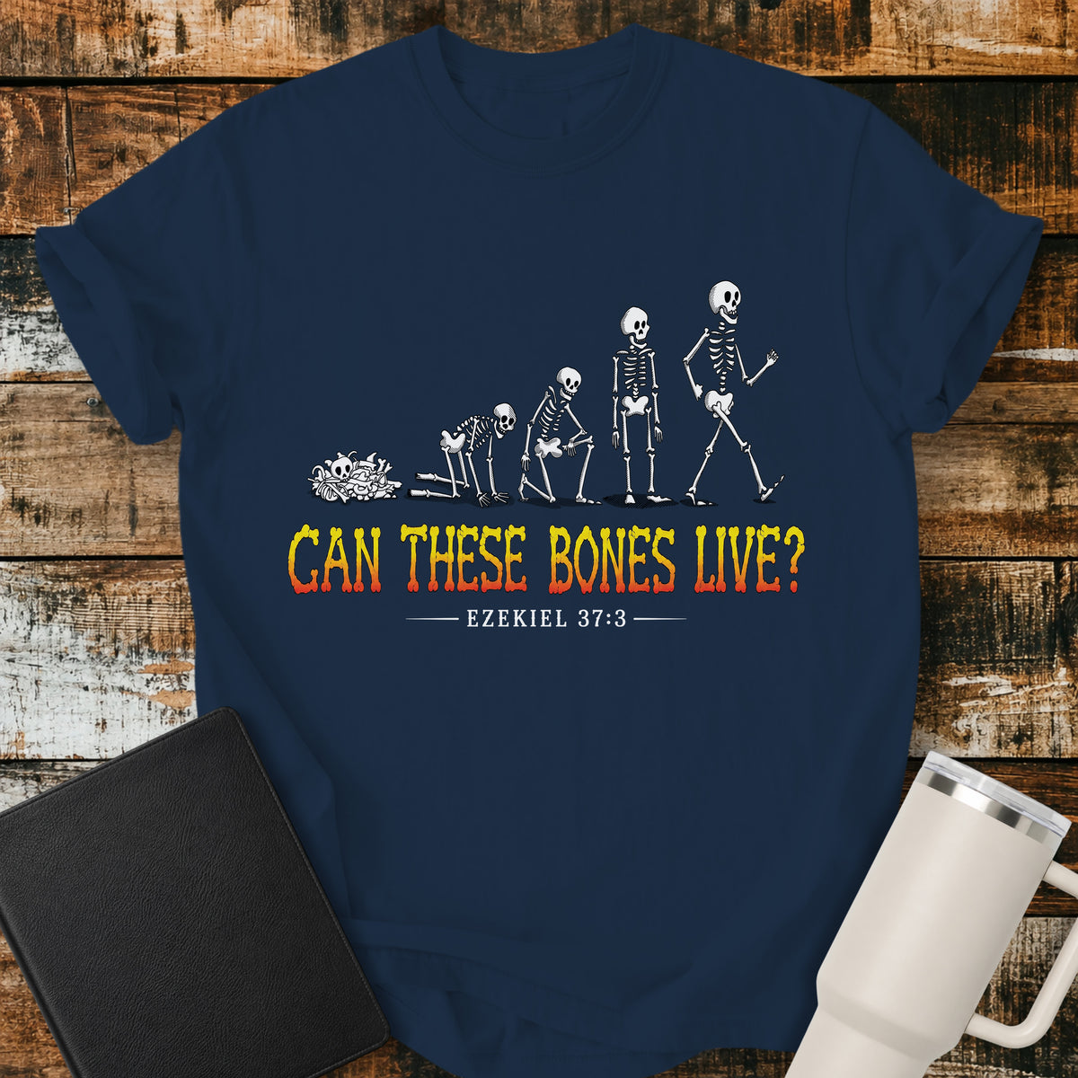 true navy flat-lay t-shirt mockup featuring a striking illustration of dry bones transforming into a walking skeleton, inspired by Ezekiel's vision of the valley of dry bones. Bold distressed lettering in a yellow-to-red gradient reads, "CAN THESE BONES LIVE?" with the Bible reference "EZEKIEL 37:3" below. The shirt is displayed on a rustic wood plank background alongside a black leather Bible and a white insulated travel mug.