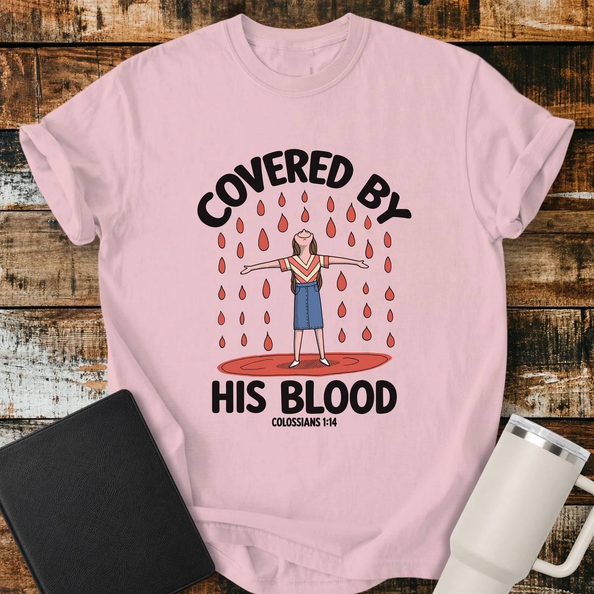 blossom pink Christian t-shirt featuring a woman standing beneath falling drops with the bold message “Covered By His Blood,” inspired by Colossians 1:14.