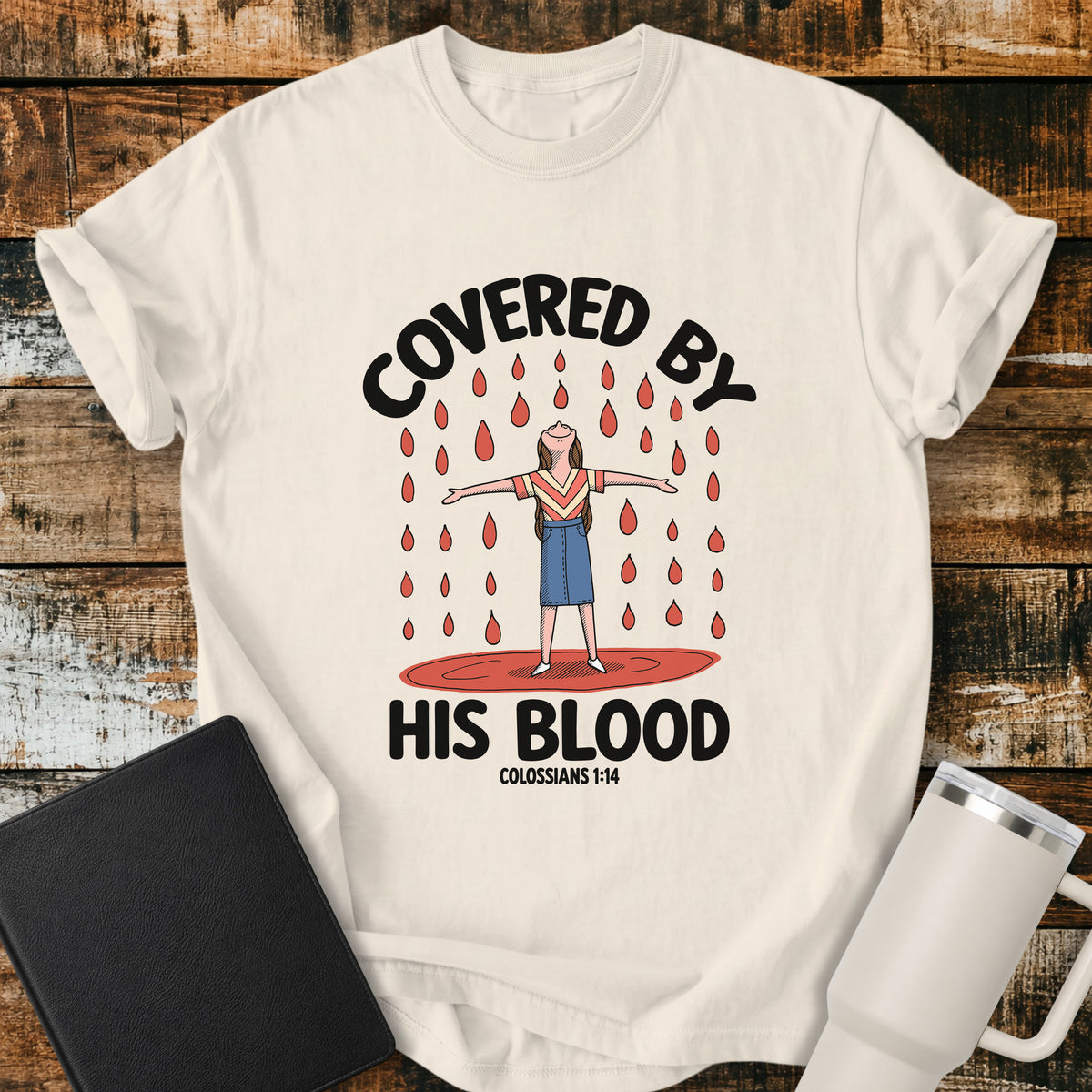 ivory Christian t-shirt featuring a woman standing beneath falling drops with the bold message “Covered By His Blood,” inspired by Colossians 1:14.