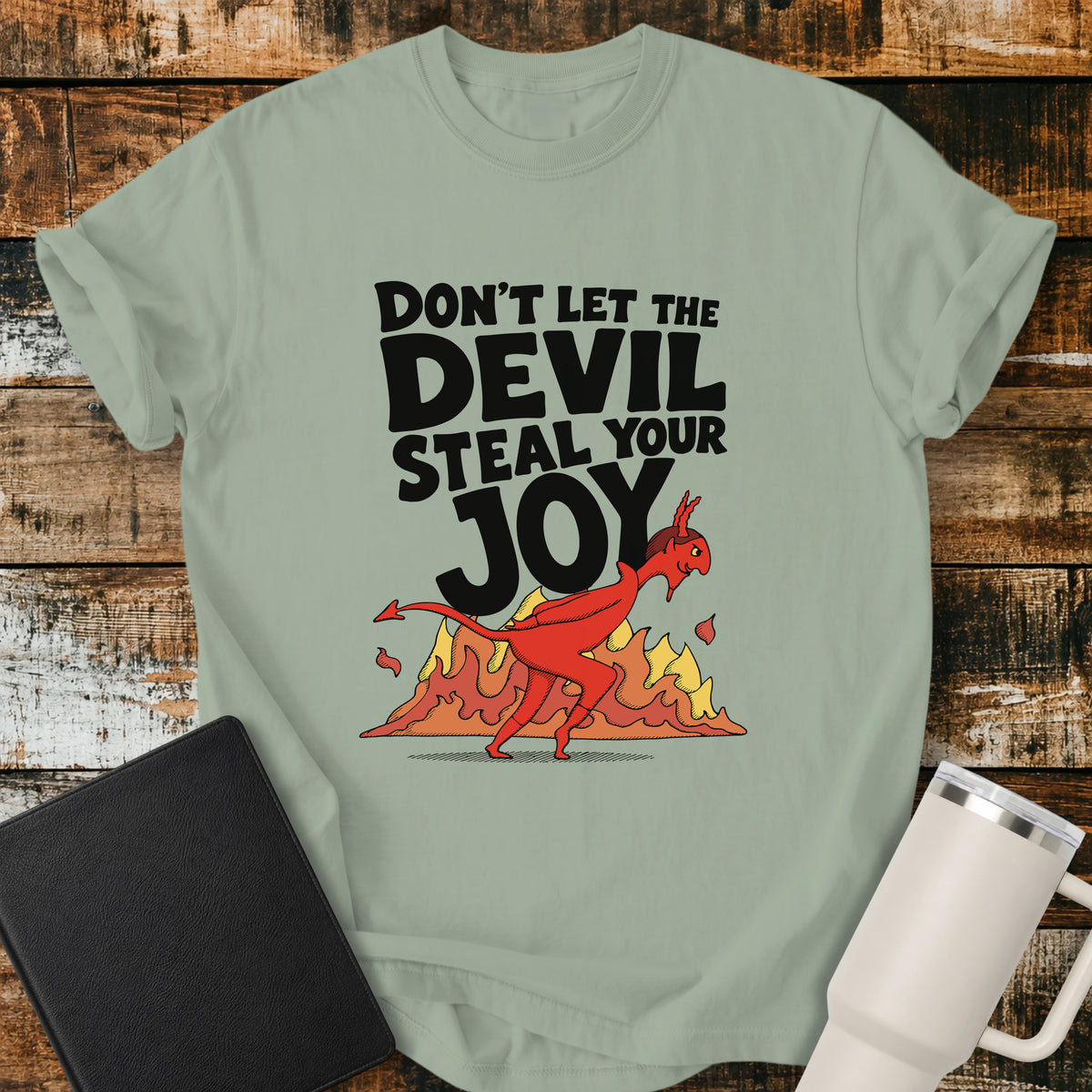 bay green Christian t-shirt featuring the bold phrase “Don’t Let the Devil Steal Your Joy” with a retro red devil sneaking away in front of stylized flames. A playful vintage-inspired faith design with bold black typography.