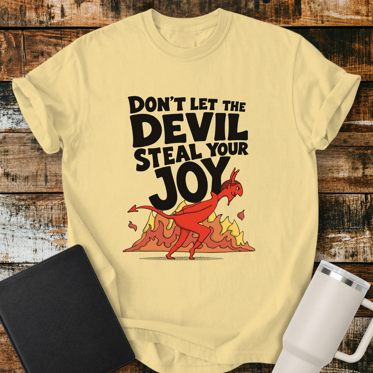Butter yellow Christian t-shirt featuring the bold phrase “Don’t Let the Devil Steal Your Joy” with a retro red devil sneaking away in front of stylized flames. A playful vintage-inspired faith design with bold black typography.