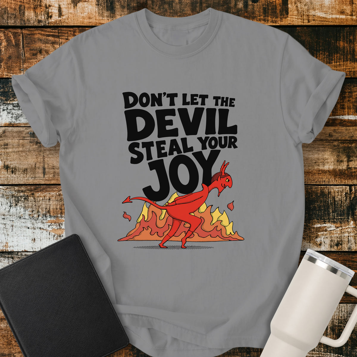 grey Christian t-shirt featuring the bold phrase “Don’t Let the Devil Steal Your Joy” with a retro red devil sneaking away in front of stylized flames. A playful vintage-inspired faith design with bold black typography.