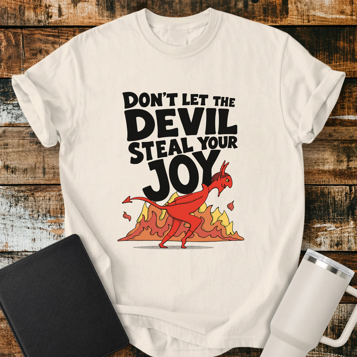 ivory Christian t-shirt featuring the bold phrase “Don’t Let the Devil Steal Your Joy” with a retro red devil sneaking away in front of stylized flames. A playful vintage-inspired faith design with bold black typography.