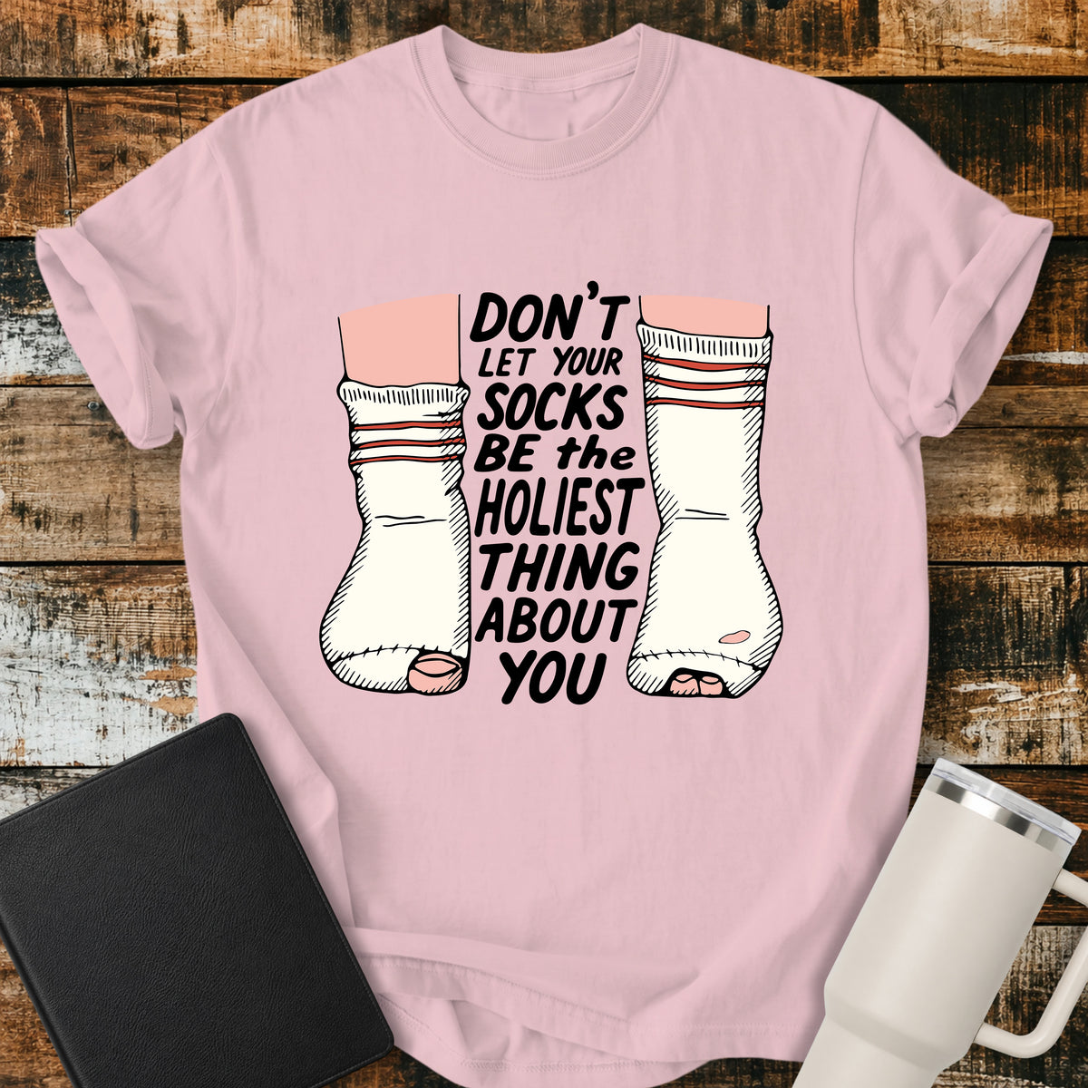 blossom pink Christian T-shirt featuring worn, holey socks with the humorous phrase “Don’t Let Your Socks Be the Holiest Thing About You” in black lettering