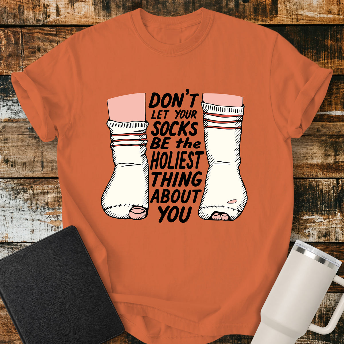 burnt orange Christian T-shirt featuring worn, holey socks with the humorous phrase “Don’t Let Your Socks Be the Holiest Thing About You” in black lettering