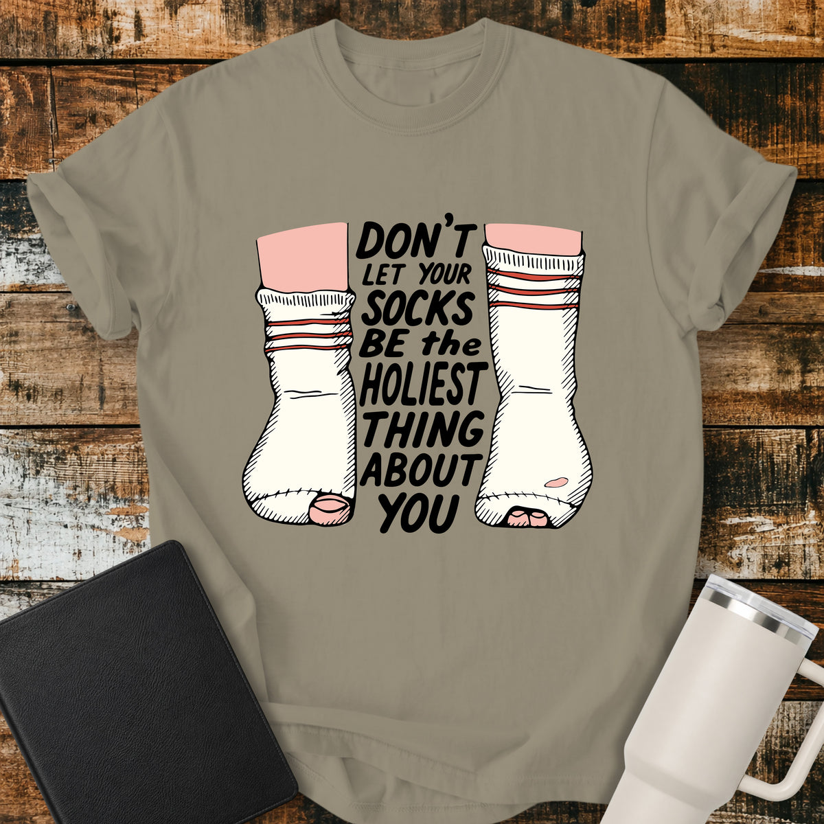 sandstone Christian T-shirt featuring worn, holey socks with the humorous phrase “Don’t Let Your Socks Be the Holiest Thing About You” in black lettering