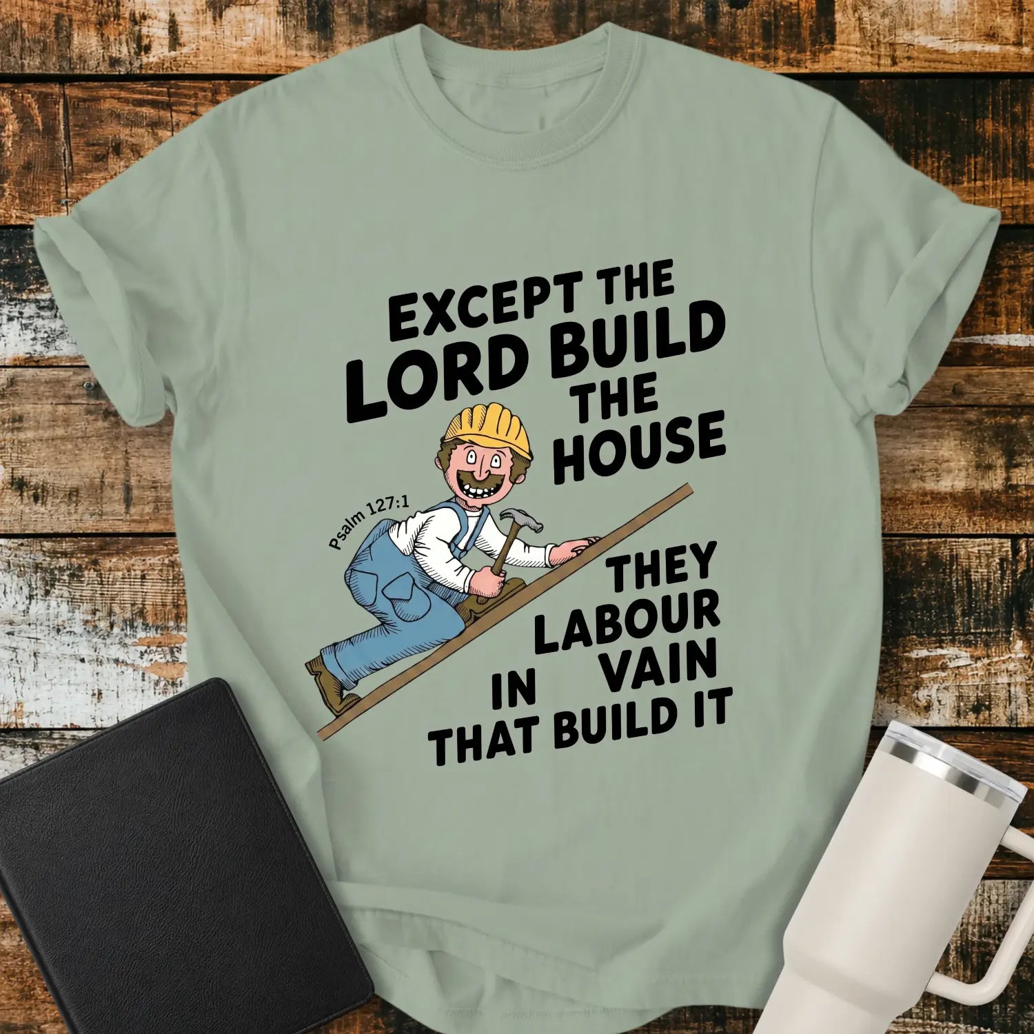 Bay green graphic T-shirt mockup featuring the Bible verse “Except the Lord Build the House, They Labour in Vain That Build It” with a Psalm 127:1 reference and a cartoon construction worker climbing a board with a hammer, styled with a black Bible and white tumbler on a rustic wood background.
