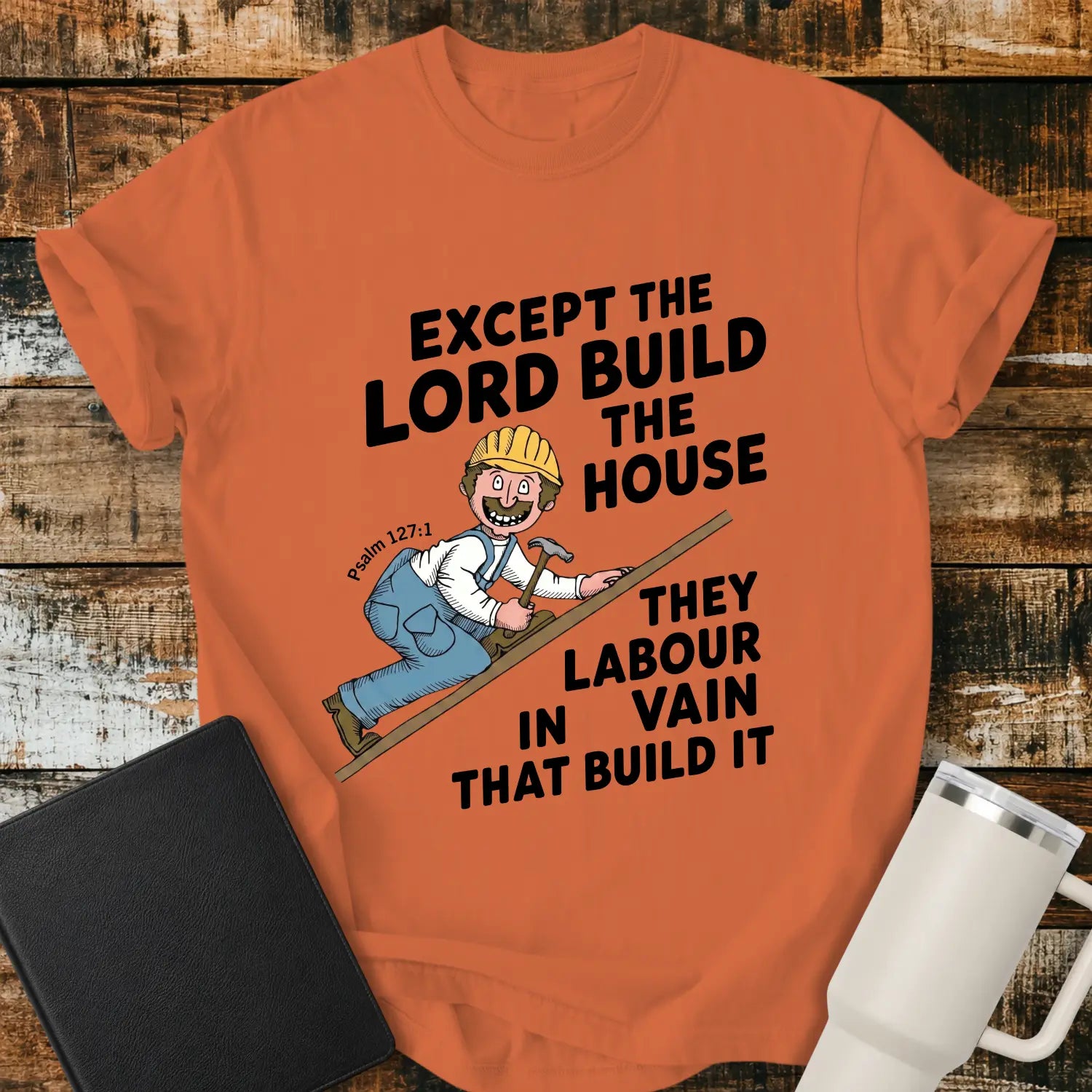 burnt orange graphic T-shirt mockup featuring the Bible verse “Except the Lord Build the House, They Labour in Vain That Build It” with a Psalm 127:1 reference and a cartoon construction worker climbing a board with a hammer, styled with a black Bible and white tumbler on a rustic wood background.
