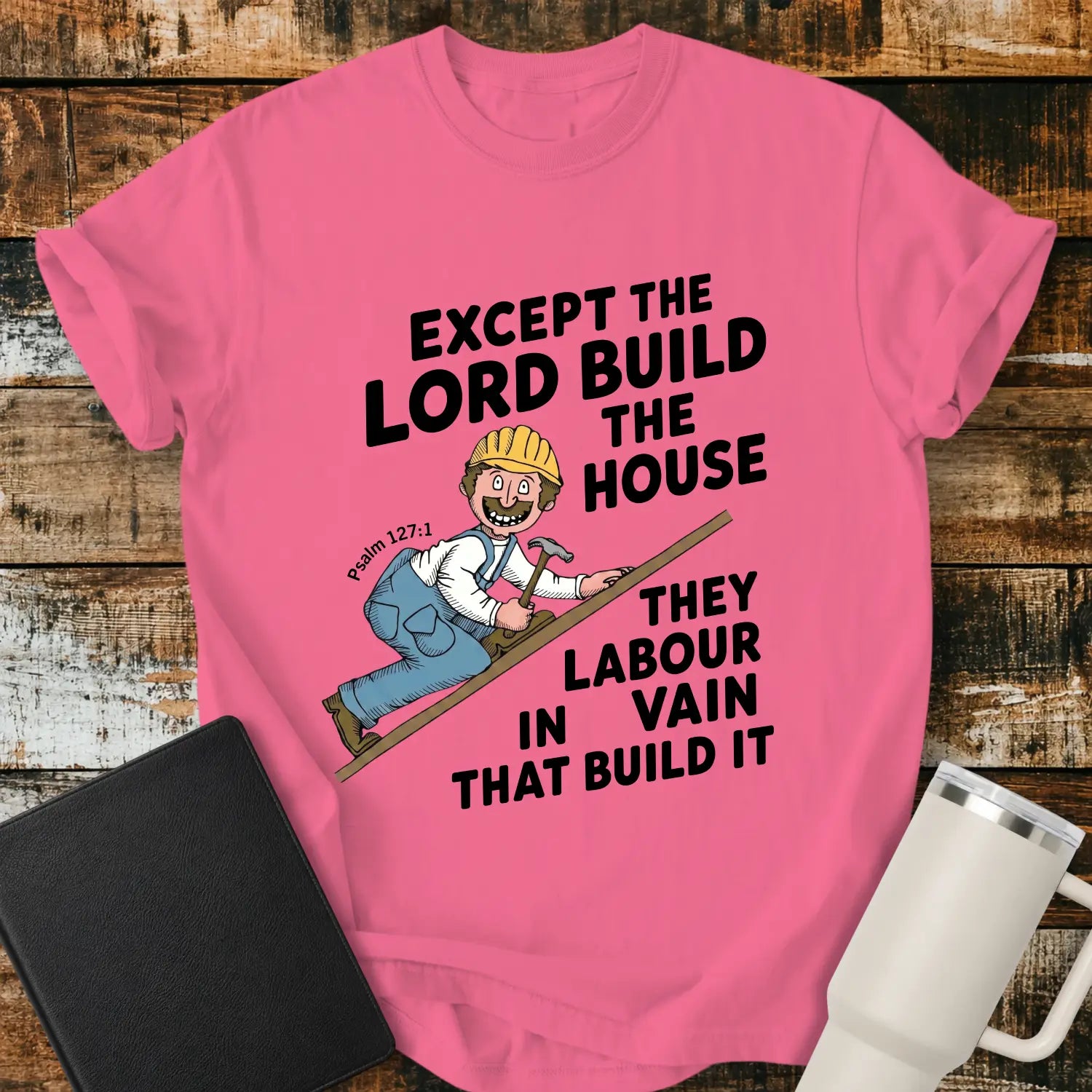 crunchberry pink graphic T-shirt mockup featuring the Bible verse “Except the Lord Build the House, They Labour in Vain That Build It” with a Psalm 127:1 reference and a cartoon construction worker climbing a board with a hammer, styled with a black Bible and white tumbler on a rustic wood background.
