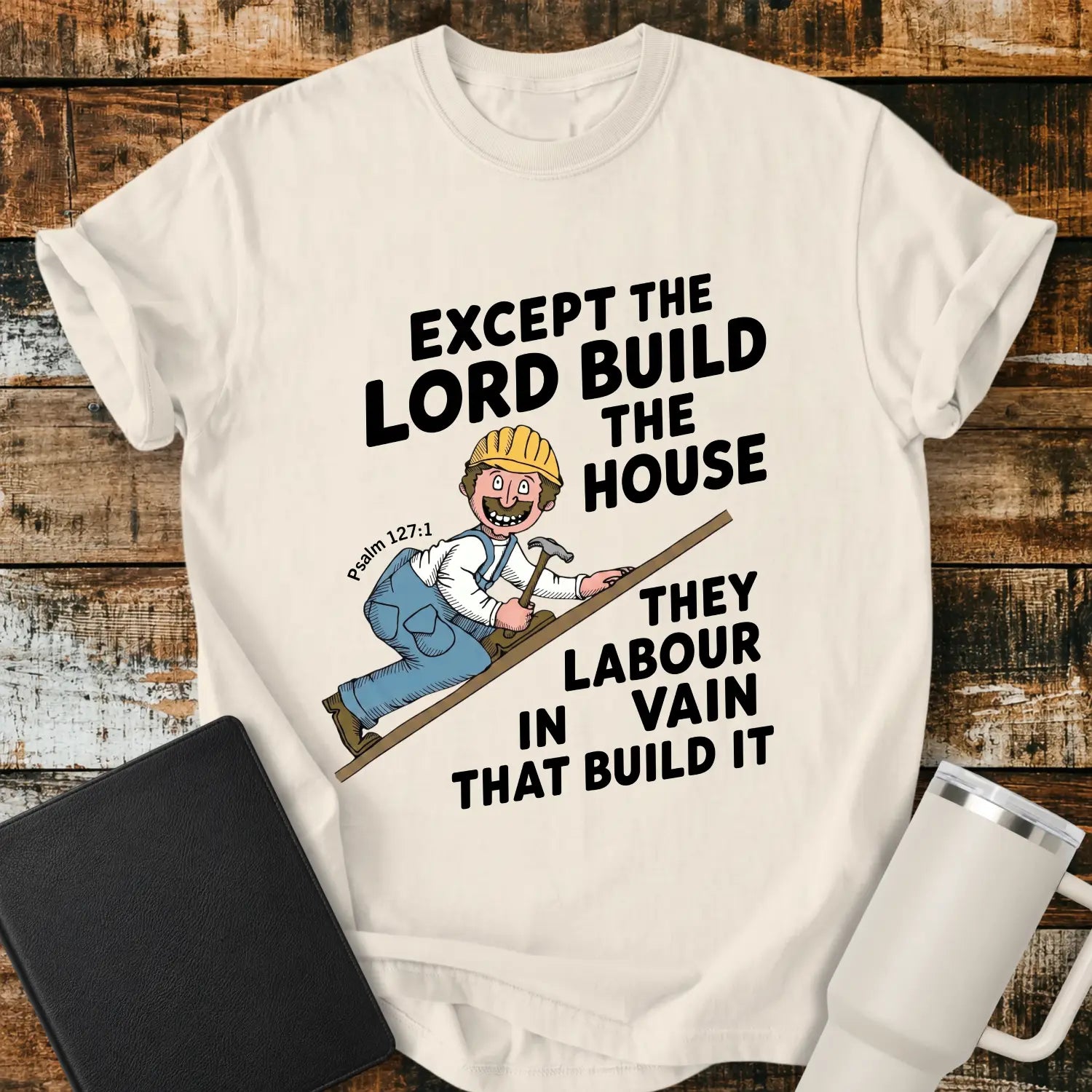 ivory graphic T-shirt mockup featuring the Bible verse “Except the Lord Build the House, They Labour in Vain That Build It” with a Psalm 127:1 reference and a cartoon construction worker climbing a board with a hammer, styled with a black Bible and white tumbler on a rustic wood background.
