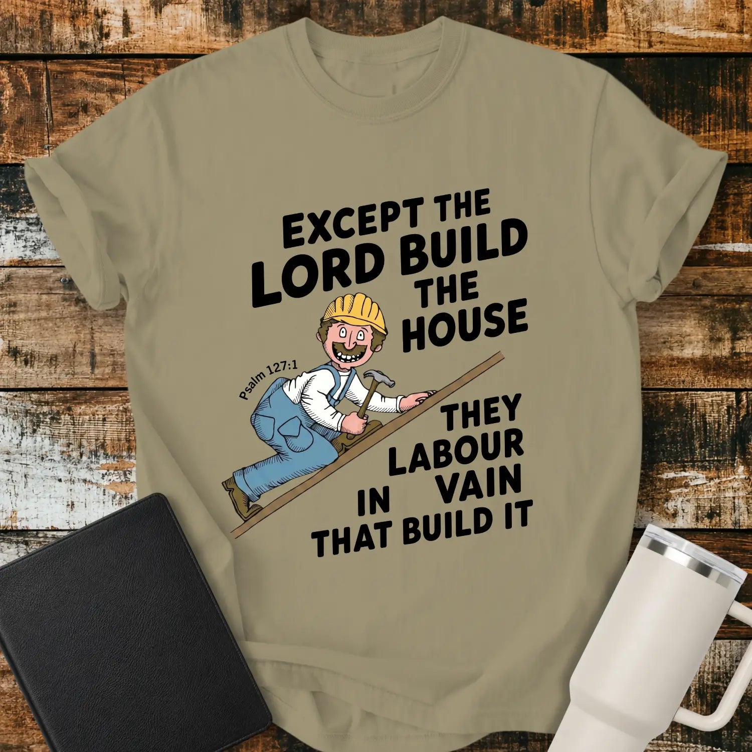 khaki graphic T-shirt mockup featuring the Bible verse “Except the Lord Build the House, They Labour in Vain That Build It” with a Psalm 127:1 reference and a cartoon construction worker climbing a board with a hammer, styled with a black Bible and white tumbler on a rustic wood background.
