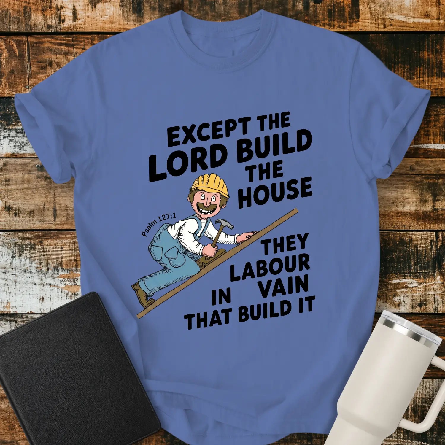 mystic blue graphic T-shirt mockup featuring the Bible verse “Except the Lord Build the House, They Labour in Vain That Build It” with a Psalm 127:1 reference and a cartoon construction worker climbing a board with a hammer, styled with a black Bible and white tumbler on a rustic wood background.
