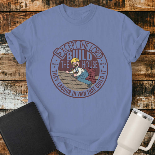 washed denim T-shirt featuring a circular vintage-inspired design based on Psalm 127:1. At the center is a smiling construction worker laying roof shingles with a stack of bricks behind him. Surrounding the illustration is the verse, “EXCEPT THE LORD BUILD THE HOUSE, THEY LABOUR IN VAIN THAT BUILD IT,” with “PSALM 127:1” incorporated into the design. The shirt is displayed on a rustic wood plank background with a black Bible on the left and a white insulated tumbler on the right.