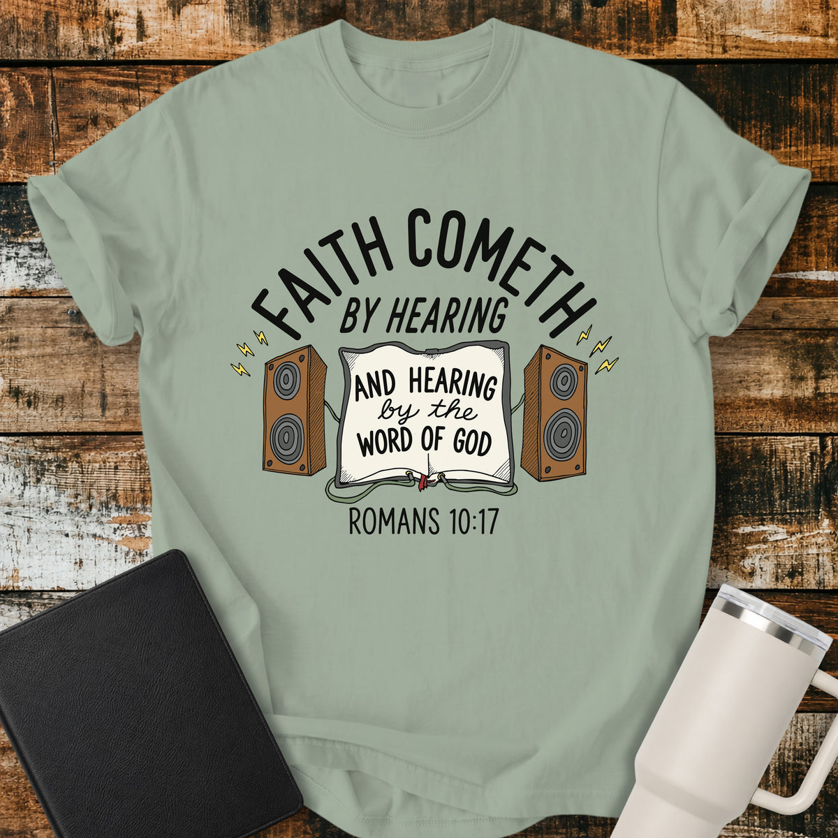 bay green Christian t-shirt featuring an open Bible connected to two speakers, with the text “Faith Cometh by Hearing and Hearing by the Word of God” and Romans 10:17. Displayed on a rustic wood background with a black Bible and white travel mug.