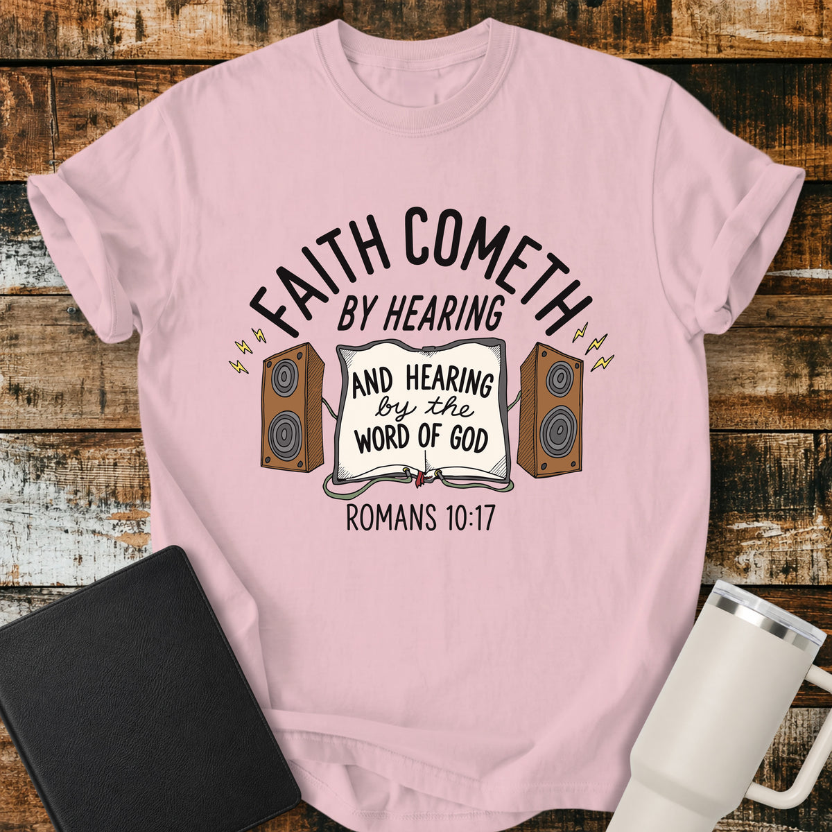 blossom pink Christian t-shirt featuring an open Bible connected to two speakers, with the text “Faith Cometh by Hearing and Hearing by the Word of God” and Romans 10:17. Displayed on a rustic wood background with a black Bible and white travel mug.