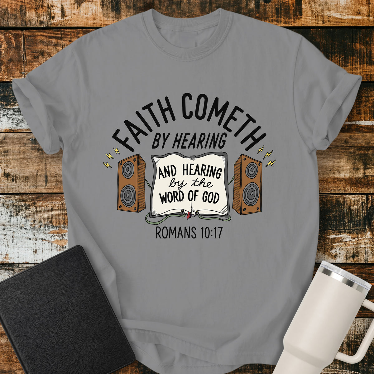 grey Christian t-shirt featuring an open Bible connected to two speakers, with the text “Faith Cometh by Hearing and Hearing by the Word of God” and Romans 10:17. Displayed on a rustic wood background with a black Bible and white travel mug.