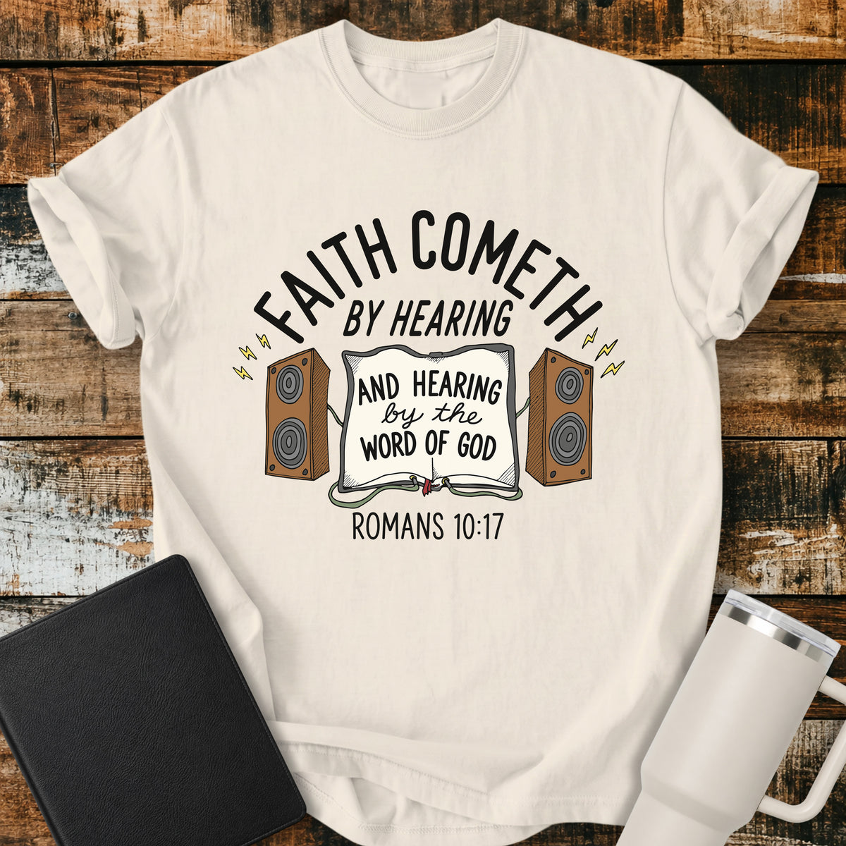 ivory Christian t-shirt featuring an open Bible connected to two speakers, with the text “Faith Cometh by Hearing and Hearing by the Word of God” and Romans 10:17. Displayed on a rustic wood background with a black Bible and white travel mug.