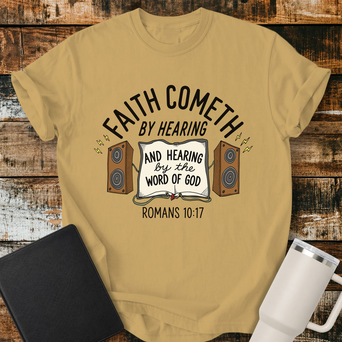 Mustard yellow Christian t-shirt featuring an open Bible connected to two speakers, with the text “Faith Cometh by Hearing and Hearing by the Word of God” and Romans 10:17. Displayed on a rustic wood background with a black Bible and white travel mug.