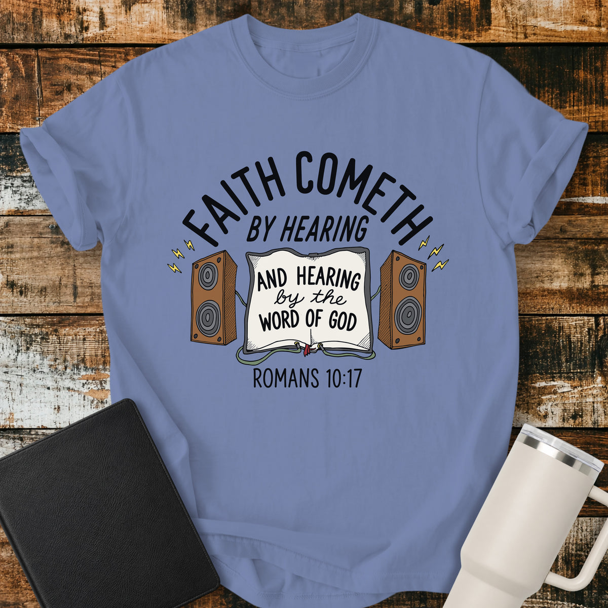 washed denim Christian t-shirt featuring an open Bible connected to two speakers, with the text “Faith Cometh by Hearing and Hearing by the Word of God” and Romans 10:17. Displayed on a rustic wood background with a black Bible and white travel mug.