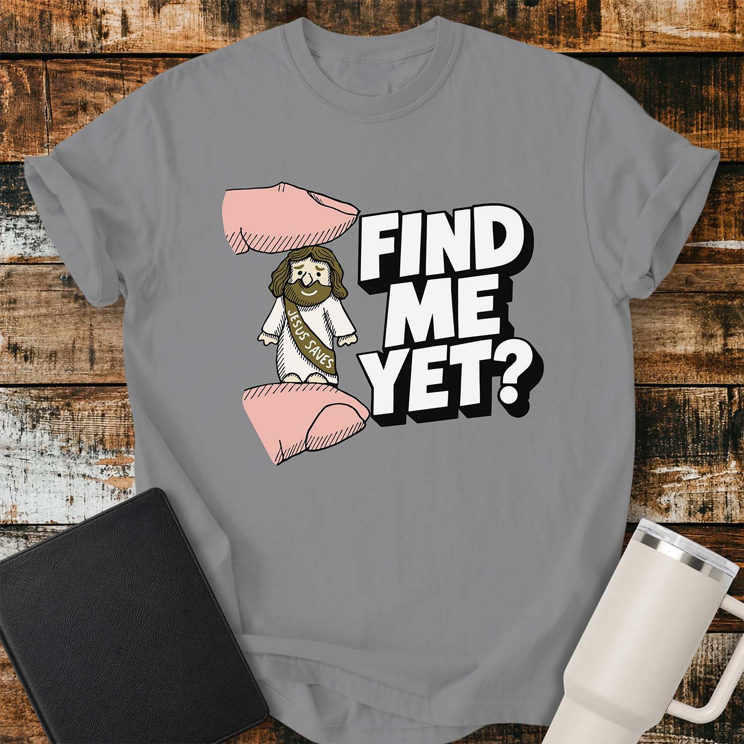 grey flat-lay t-shirt mockup featuring a tiny Jesus figurine standing between two fingers as if being pinched, alongside bold white block lettering that reads, "FIND ME YET?" The figurine wears a sash that says "JESUS SAVES," emphasizing its miniature size. The shirt is displayed on a rustic wood plank background with a black leather Bible and a white insulated travel mug.