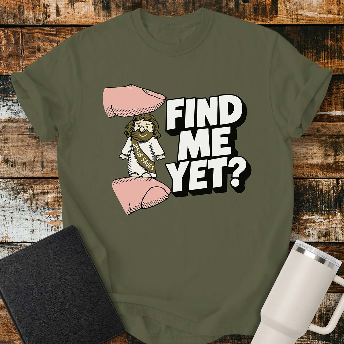 hemp green flat-lay t-shirt mockup featuring a tiny Jesus figurine standing between two fingers as if being pinched, alongside bold white block lettering that reads, "FIND ME YET?" The figurine wears a sash that says "JESUS SAVES," emphasizing its miniature size. The shirt is displayed on a rustic wood plank background with a black leather Bible and a white insulated travel mug.