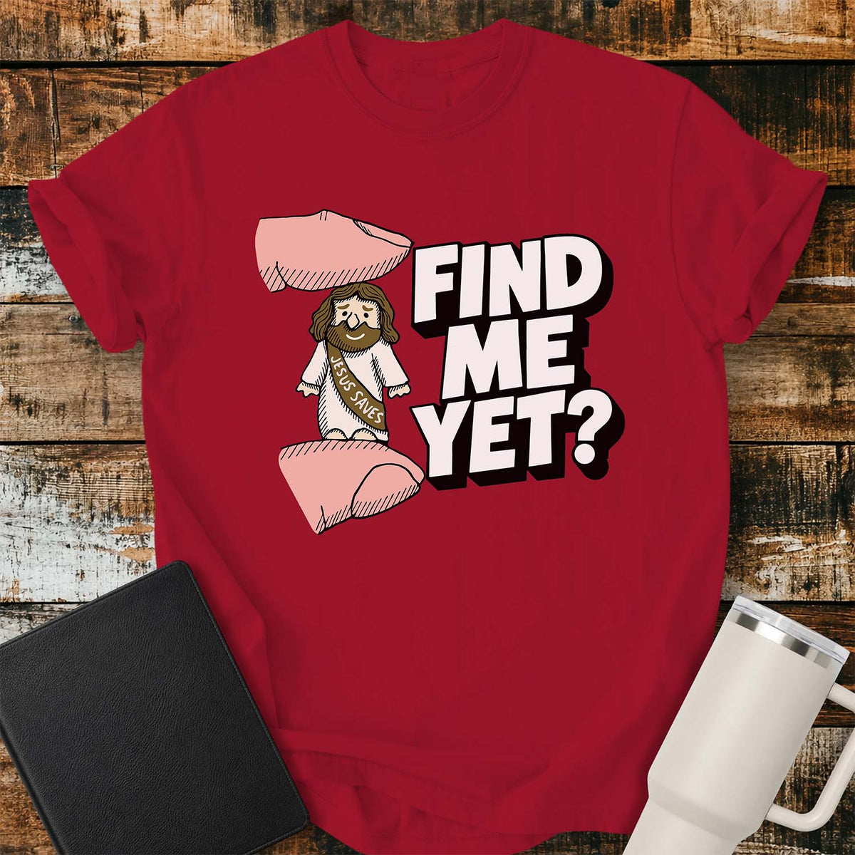 red flat-lay t-shirt mockup featuring a tiny Jesus figurine standing between two fingers as if being pinched, alongside bold white block lettering that reads, "FIND ME YET?" The figurine wears a sash that says "JESUS SAVES," emphasizing its miniature size. The shirt is displayed on a rustic wood plank background with a black leather Bible and a white insulated travel mug.