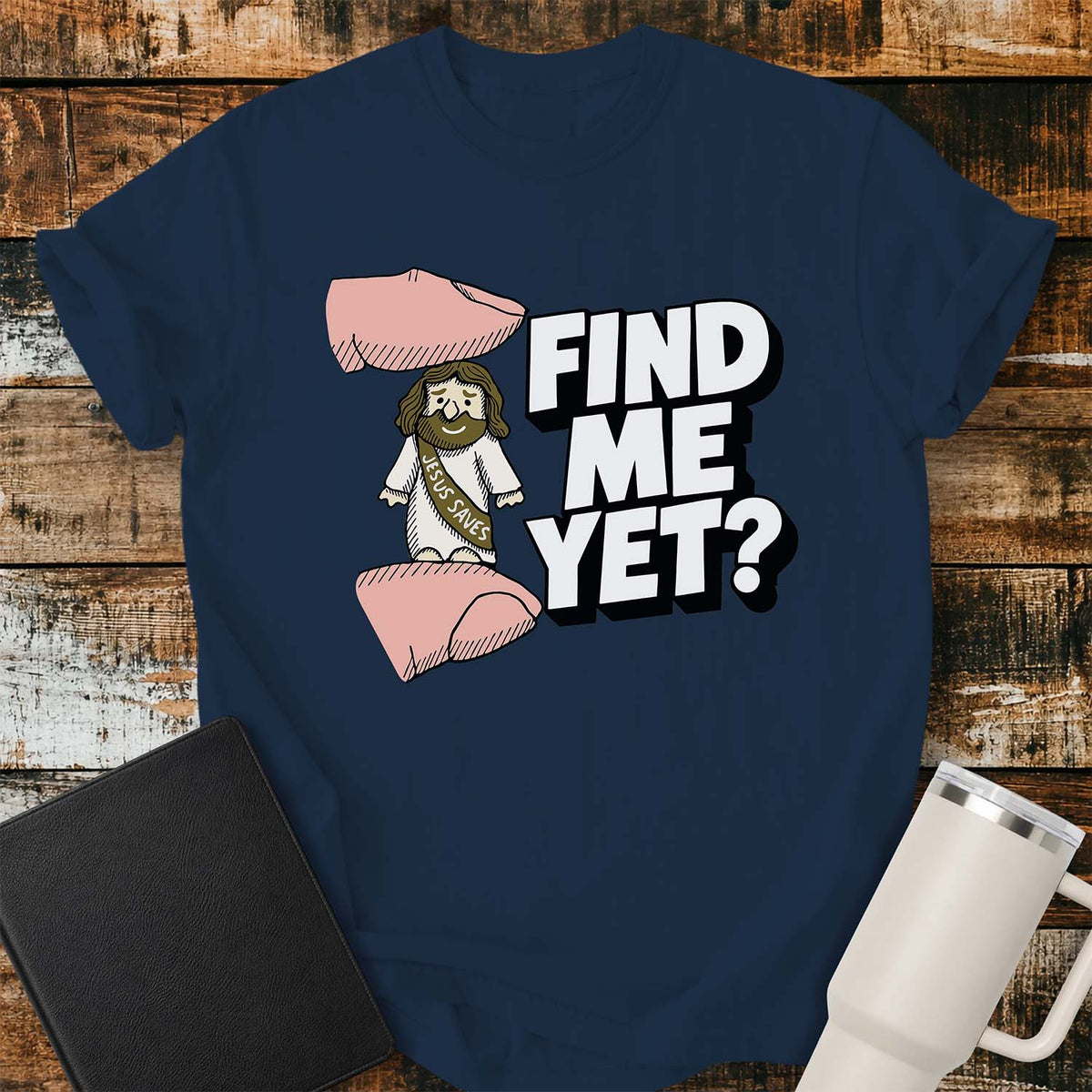 true navy flat-lay t-shirt mockup featuring a tiny Jesus figurine standing between two fingers as if being pinched, alongside bold white block lettering that reads, "FIND ME YET?" The figurine wears a sash that says "JESUS SAVES," emphasizing its miniature size. The shirt is displayed on a rustic wood plank background with a black leather Bible and a white insulated travel mug.