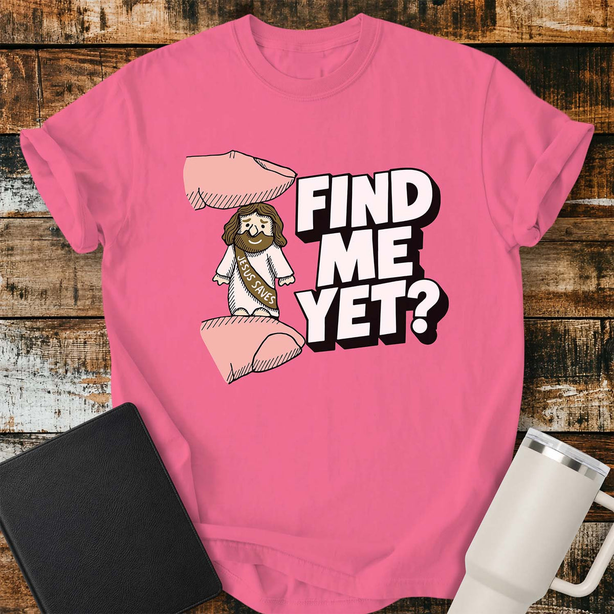 crunchberry pink flat-lay t-shirt mockup featuring a tiny Jesus figurine standing between two fingers as if being pinched, alongside bold white block lettering that reads, "FIND ME YET?" The figurine wears a sash that says "JESUS SAVES," emphasizing its miniature size. The shirt is displayed on a rustic wood plank background with a black leather Bible and a white insulated travel mug.