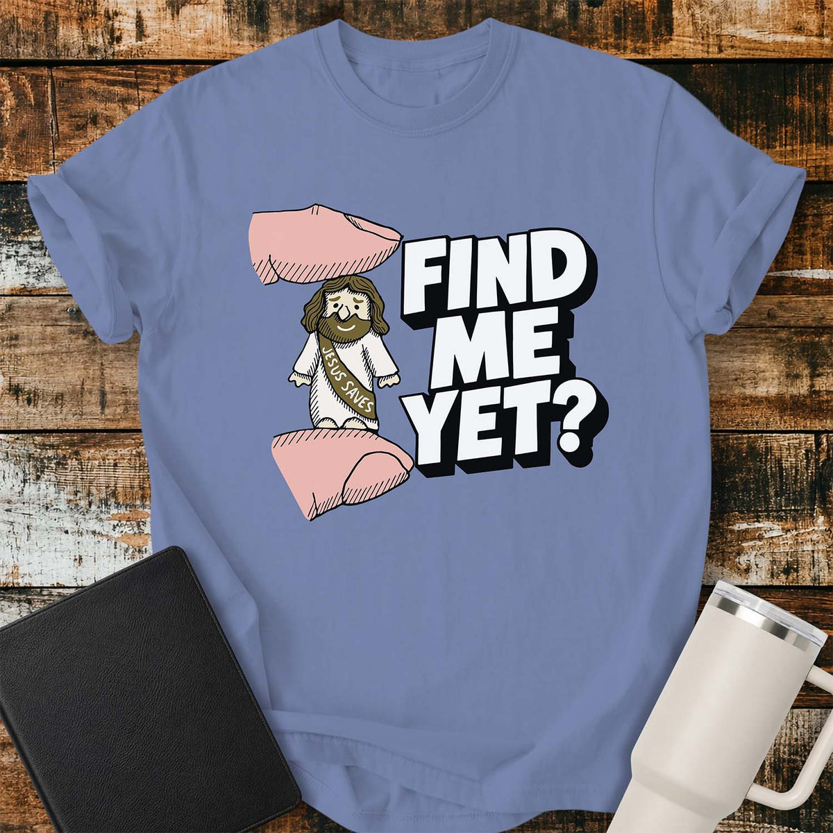 washed denim flat-lay t-shirt mockup featuring a tiny Jesus figurine standing between two fingers as if being pinched, alongside bold white block lettering that reads, "FIND ME YET?" The figurine wears a sash that says "JESUS SAVES," emphasizing its miniature size. The shirt is displayed on a rustic wood plank background with a black leather Bible and a white insulated travel mug.