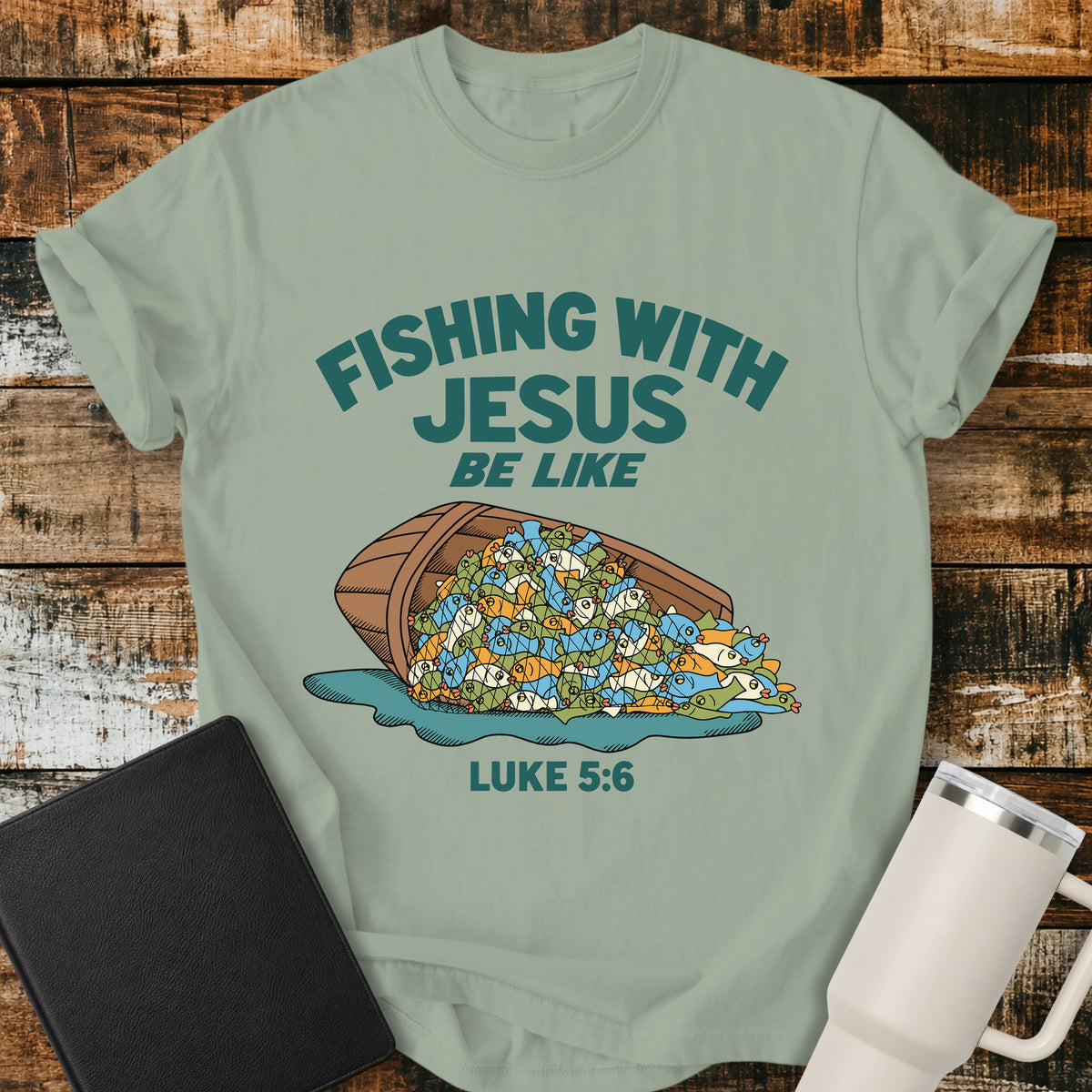 bay green Christian t-shirt featuring a humorous overflowing catch of fish spilling from a boat, with bold retro text reading “Fishing With Jesus Be Like” and Luke 5:6.