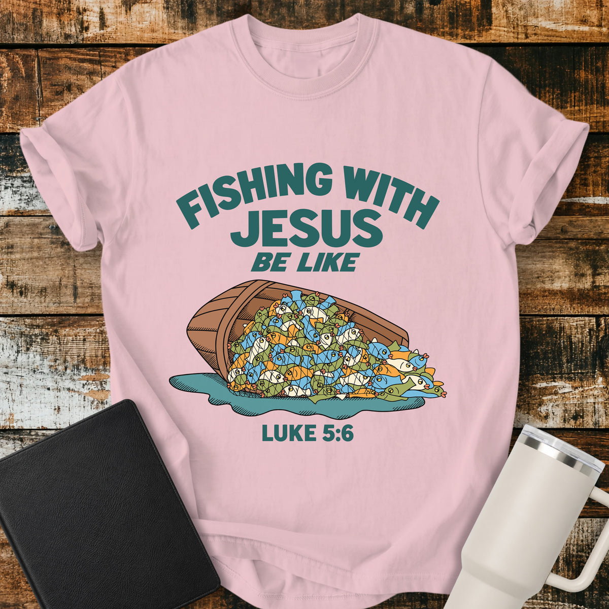 blossom pink Christian t-shirt featuring a humorous overflowing catch of fish spilling from a boat, with bold retro text reading “Fishing With Jesus Be Like” and Luke 5:6.