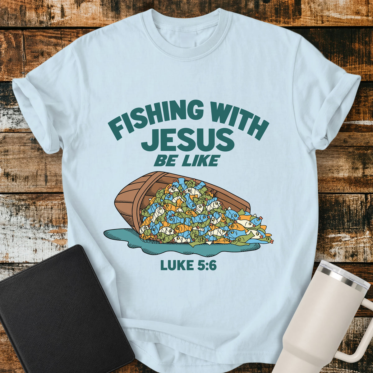 chambray blue Christian t-shirt featuring a humorous overflowing catch of fish spilling from a boat, with bold retro text reading “Fishing With Jesus Be Like” and Luke 5:6.