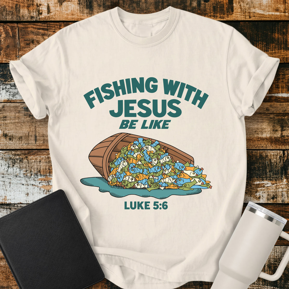 Ivory Christian t-shirt featuring a humorous overflowing catch of fish spilling from a boat, with bold retro text reading “Fishing With Jesus Be Like” and Luke 5:6.