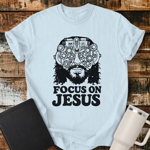 Chambray blue T-shirt featuring a black vintage-style illustration of Jesus wearing a detailed phoropter over His eyes with the bold phrase “FOCUS ON JESUS” beneath. The design has a retro, hand-drawn aesthetic with thick black lettering. The shirt is displayed on a rustic wood plank background with a black Bible on the left and a white insulated tumbler on the right.