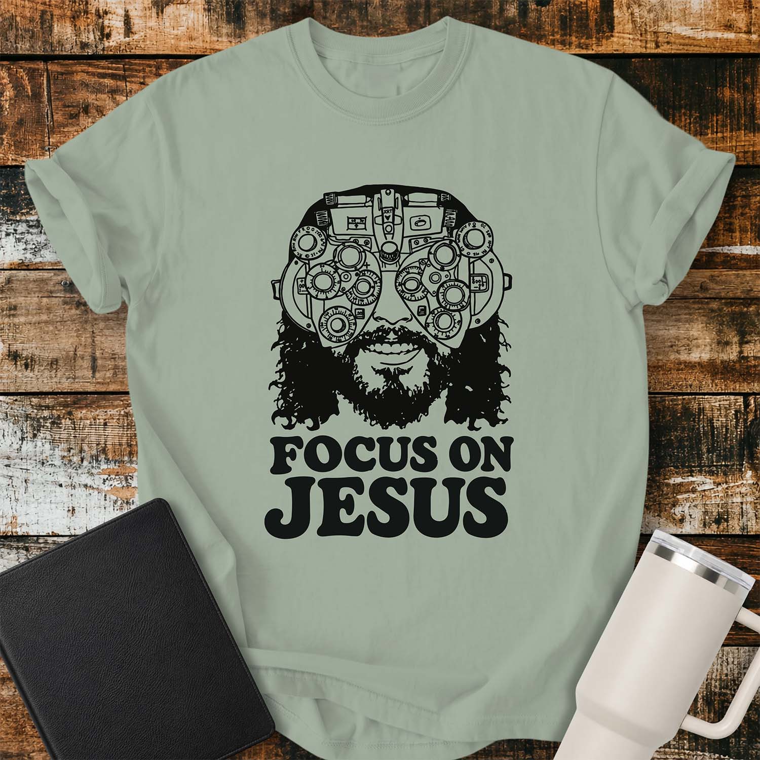 Bay green T-shirt featuring a black vintage-style illustration of Jesus wearing a detailed phoropter over His eyes with the bold phrase “FOCUS ON JESUS” beneath. The design has a retro, hand-drawn aesthetic with thick black lettering. The shirt is displayed on a rustic wood plank background with a black Bible on the left and a white insulated tumbler on the right.