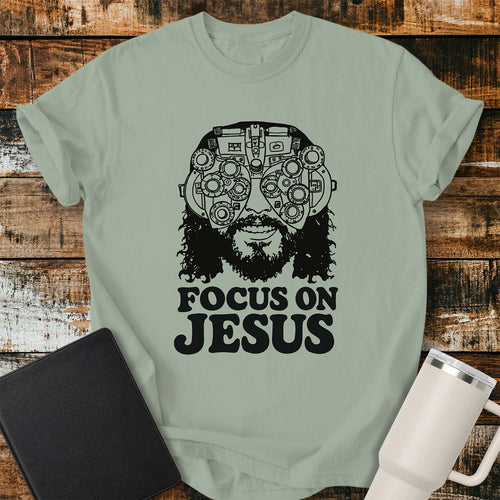 Bay green T-shirt featuring a black vintage-style illustration of Jesus wearing a detailed phoropter over His eyes with the bold phrase “FOCUS ON JESUS” beneath. The design has a retro, hand-drawn aesthetic with thick black lettering. The shirt is displayed on a rustic wood plank background with a black Bible on the left and a white insulated tumbler on the right.