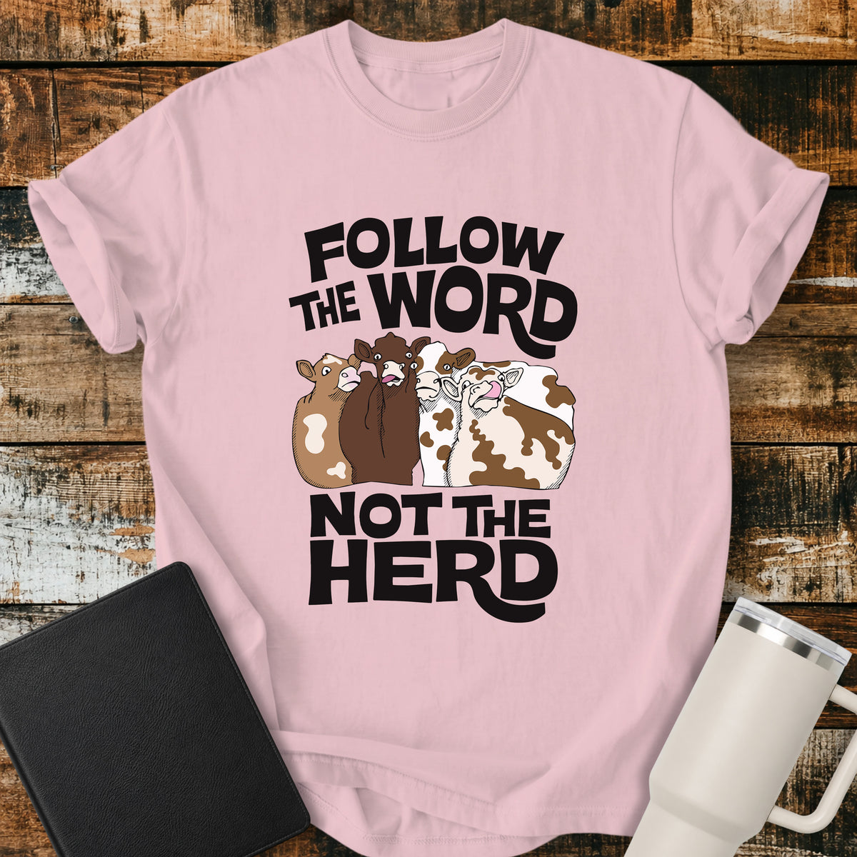 blossom pink flat-lay t-shirt mockup featuring four cartoon cows standing together beneath bold retro black lettering that reads, "FOLLOW THE WORD" above and "NOT THE HERD" below. The faith-inspired design encourages following God's Word rather than the crowd. The shirt is displayed on a rustic wood plank background alongside a black leather Bible and a white insulated travel mug.