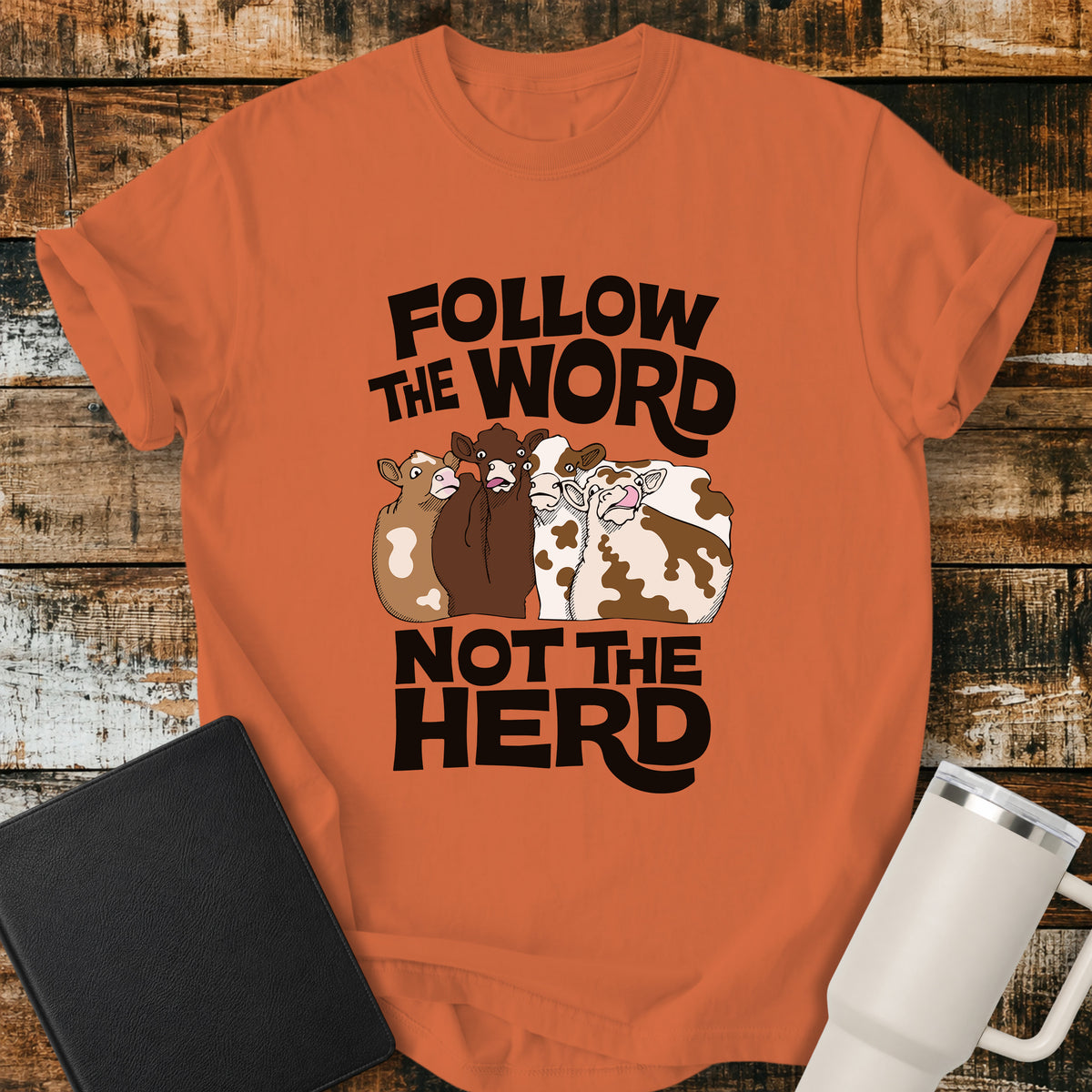 burnt orange flat-lay t-shirt mockup featuring four cartoon cows standing together beneath bold retro black lettering that reads, "FOLLOW THE WORD" above and "NOT THE HERD" below. The faith-inspired design encourages following God's Word rather than the crowd. The shirt is displayed on a rustic wood plank background alongside a black leather Bible and a white insulated travel mug.