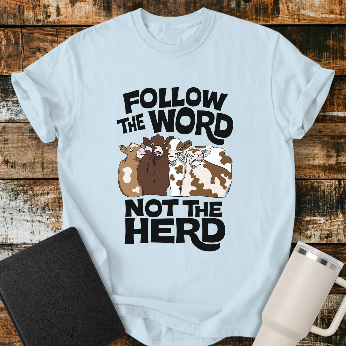 chambray blue flat-lay t-shirt mockup featuring four cartoon cows standing together beneath bold retro black lettering that reads, "FOLLOW THE WORD" above and "NOT THE HERD" below. The faith-inspired design encourages following God's Word rather than the crowd. The shirt is displayed on a rustic wood plank background alongside a black leather Bible and a white insulated travel mug.