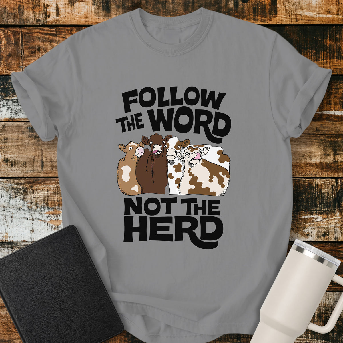 grey flat-lay t-shirt mockup featuring four cartoon cows standing together beneath bold retro black lettering that reads, "FOLLOW THE WORD" above and "NOT THE HERD" below. The faith-inspired design encourages following God's Word rather than the crowd. The shirt is displayed on a rustic wood plank background alongside a black leather Bible and a white insulated travel mug.