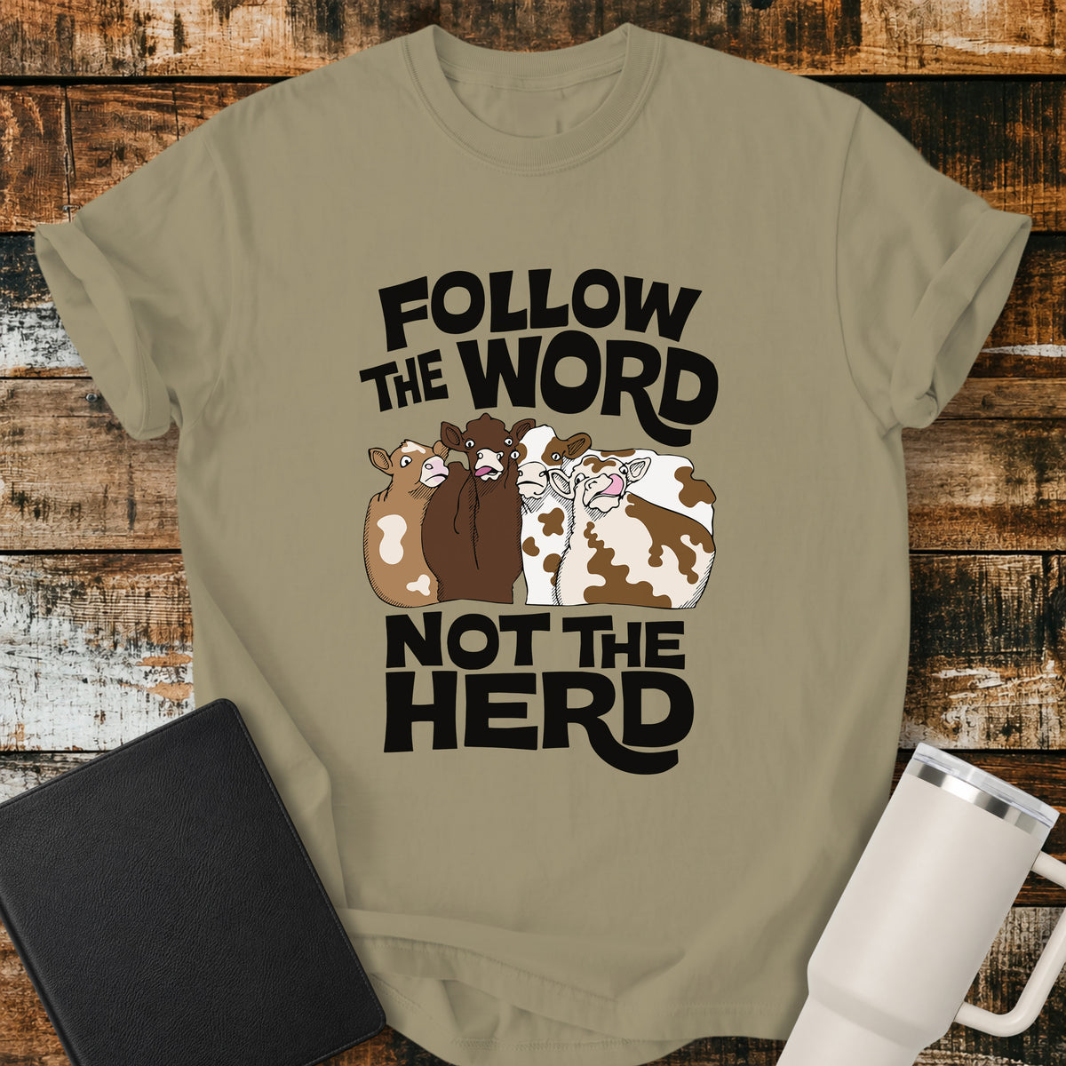 khaki flat-lay t-shirt mockup featuring four cartoon cows standing together beneath bold retro black lettering that reads, "FOLLOW THE WORD" above and "NOT THE HERD" below. The faith-inspired design encourages following God's Word rather than the crowd. The shirt is displayed on a rustic wood plank background alongside a black leather Bible and a white insulated travel mug.