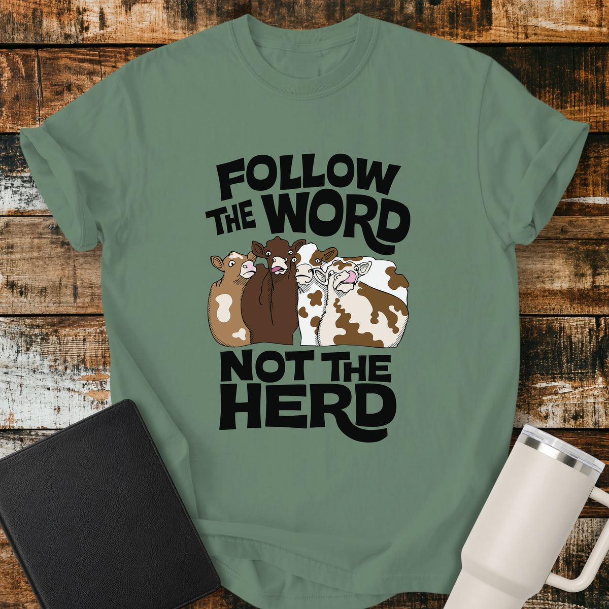light green flat-lay t-shirt mockup featuring four cartoon cows standing together beneath bold retro black lettering that reads, "FOLLOW THE WORD" above and "NOT THE HERD" below. The faith-inspired design encourages following God's Word rather than the crowd. The shirt is displayed on a rustic wood plank background alongside a black leather Bible and a white insulated travel mug.