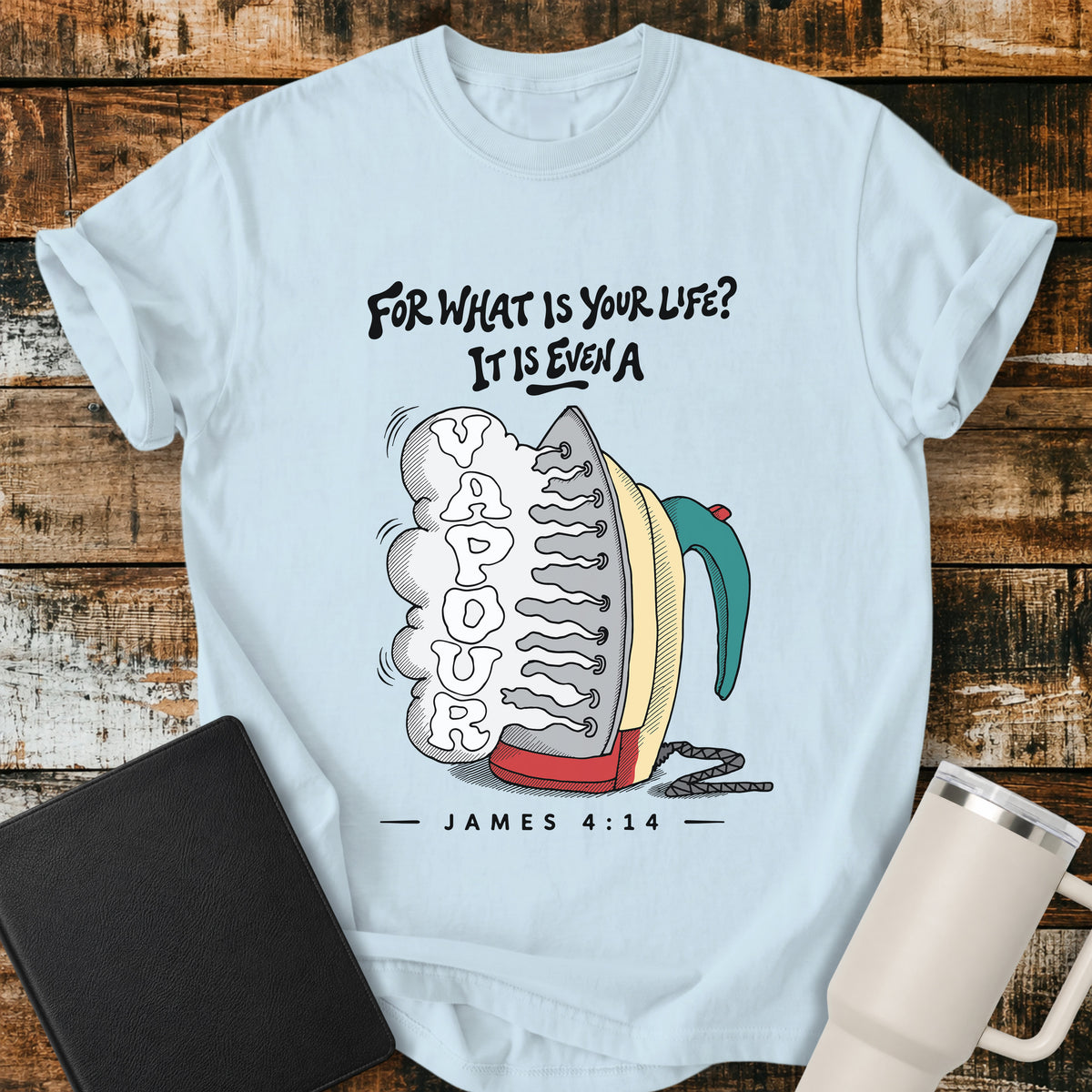 chambray blue flat-lay t-shirt mockup featuring a colorful vintage-style steam iron releasing a large cloud of steam shaped into the word "VAPOUR." Above the illustration, stylized lettering reads, "FOR WHAT IS YOUR LIFE? IT IS EVEN A," with the Bible reference "JAMES 4:14" below. The faith-inspired design is displayed on a rustic wood plank background alongside a black leather Bible and a white insulated travel mug.