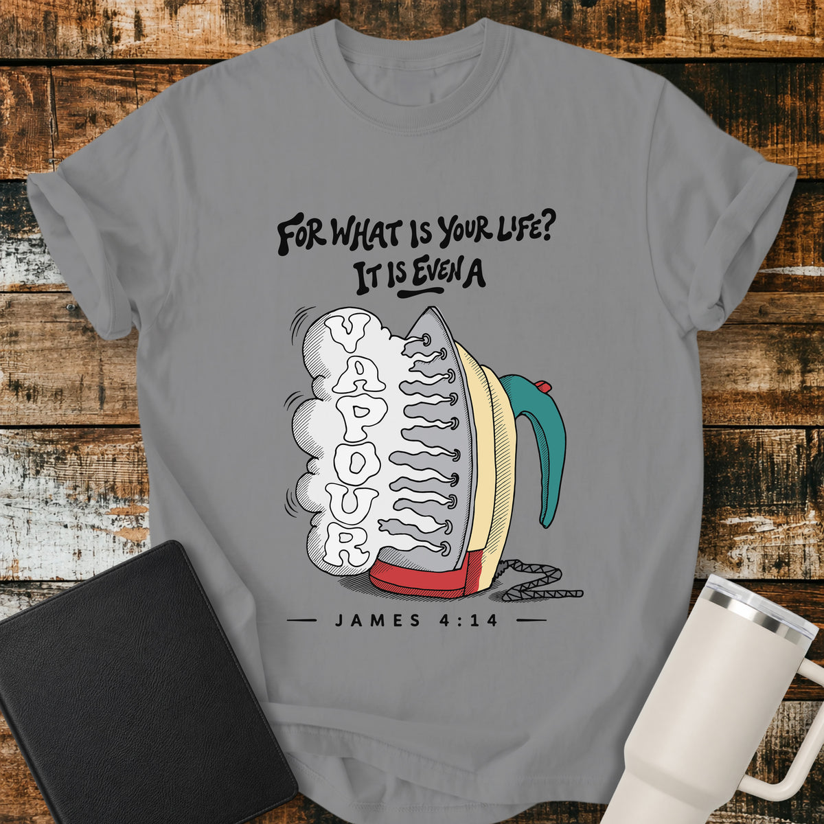 grey flat-lay t-shirt mockup featuring a colorful vintage-style steam iron releasing a large cloud of steam shaped into the word "VAPOUR." Above the illustration, stylized lettering reads, "FOR WHAT IS YOUR LIFE? IT IS EVEN A," with the Bible reference "JAMES 4:14" below. The faith-inspired design is displayed on a rustic wood plank background alongside a black leather Bible and a white insulated travel mug.