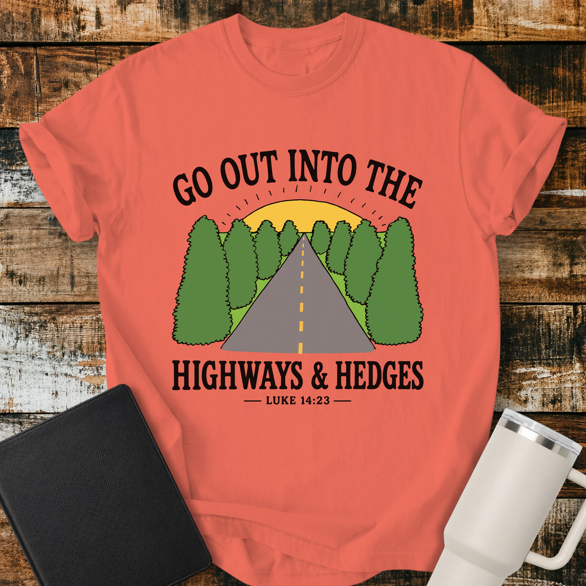 Bright salmon Christian t-shirt featuring a retro highway stretching through green hedges toward a sunrise, with the message “Go Out Into the Highways & Hedges” and the scripture reference Luke 14:23.