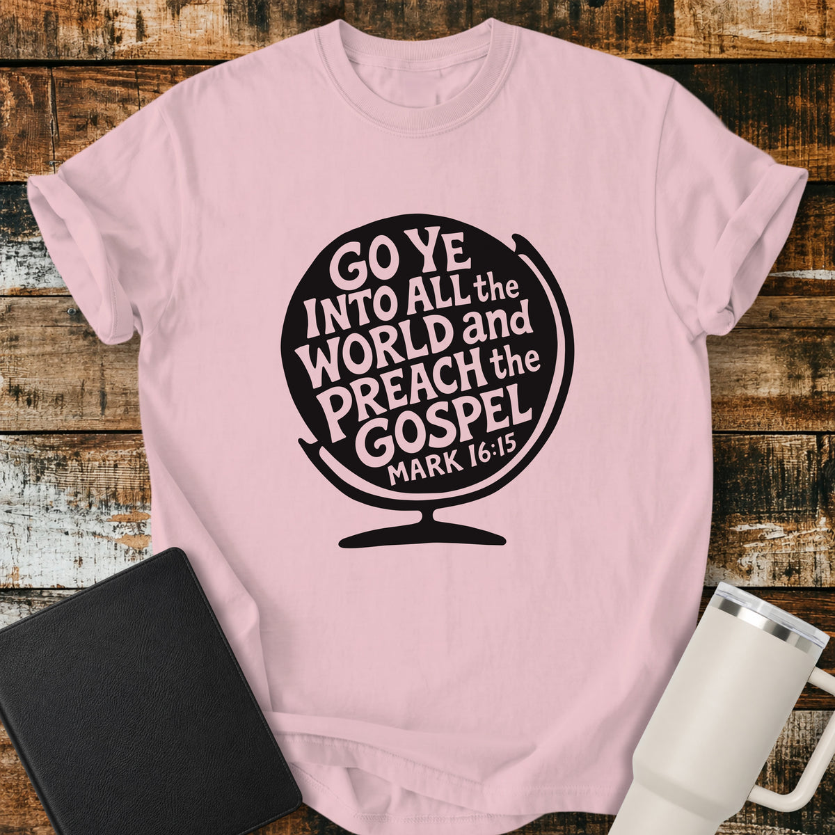 blossom pink flat-lay t-shirt mockup featuring a black globe silhouette filled with stylized lettering that reads, "GO YE INTO ALL THE WORLD AND PREACH THE GOSPEL," with the Bible reference "MARK 16:15" at the bottom. The faith-inspired design is centered on the shirt and displayed on a rustic wood plank background alongside a black leather Bible and a white insulated travel mug.