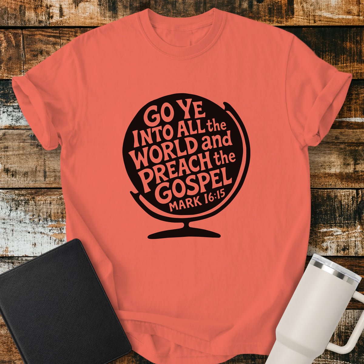 bright salmon orange flat-lay t-shirt mockup featuring a black globe silhouette filled with stylized lettering that reads, "GO YE INTO ALL THE WORLD AND PREACH THE GOSPEL," with the Bible reference "MARK 16:15" at the bottom. The faith-inspired design is centered on the shirt and displayed on a rustic wood plank background alongside a black leather Bible and a white insulated travel mug.