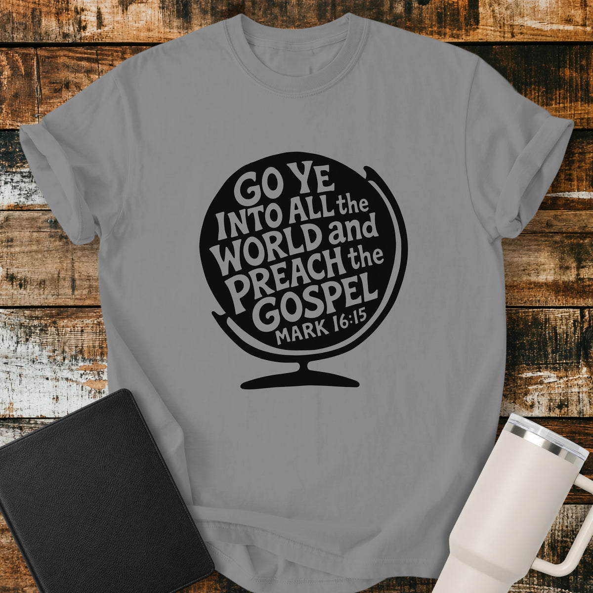 grey flat-lay t-shirt mockup featuring a black globe silhouette filled with stylized lettering that reads, "GO YE INTO ALL THE WORLD AND PREACH THE GOSPEL," with the Bible reference "MARK 16:15" at the bottom. The faith-inspired design is centered on the shirt and displayed on a rustic wood plank background alongside a black leather Bible and a white insulated travel mug.